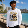 Man wearing a white long-sleeve shirt with a graphic design of a woman and text, standing on a beach with palm trees and buildings in the background.